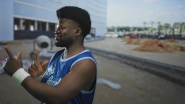 İnşaat binasında mavi basketbol forması ve bileklik takan çıplak kollu bir adam başparmak kaldırıyor..