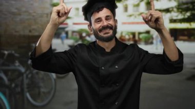 Sokak ortasında gülümseyen sakallı İspanyol erkek şef, gelişigüzel bir şehir ortamında neşeyle işaret ediyor..