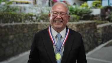 Takım elbise ve madalya giyen kıdemli bir adam sokakta el hareketi yapıyor, açık hava yeşilliğinin ortasında gülümsüyor, formalite ve mizahın bir karışımını yakalıyor..