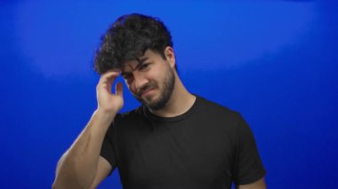 Young man with beard scratching head against vibrant blue background looking confused thoughtful contemplative expression pondering casual clothing dark hair.