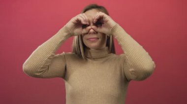 Sarışın, neşeli bir şekilde elleri ile izole edilmiş kırmızı duvar arkaplanına dürbün yapan genç bir kadın neşeli ve neşeli bir ifade sergiliyor..