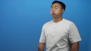 Genç adam, mavi duvarla çerçevelenmiş stüdyoda duran yanaklarını şişiriyor yarı kapalı beyaz gömlekli, can sıkıntısı.