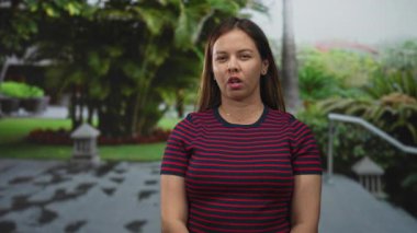 Genç bir kadın, tropik bitkiler ve taş yollarla dolu bir caddede çizgili bir tepede dudaklarını büzüyor..