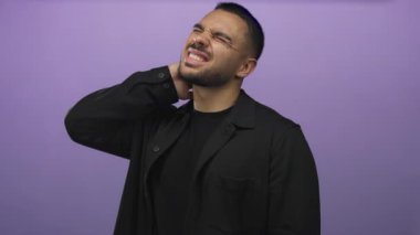 Siyah tişört giyen İspanyol genç adam mor stüdyoda boynuna dokunuyor gözleri kapalı ve gergin bir ifadeyle;.