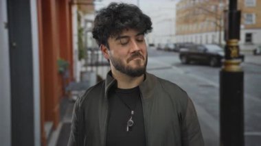 Sakallı genç adam gündüz vakti şehir sokağında duruyor, kolye ve günlük ceket giyiyor, dostça gülümsemesi ve binaların ve arabaların geçmişiyle İspanyol görünümünü sergiliyor.