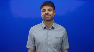 Latin kökenli genç adam gülümser ve mavi stüdyoda el hareketleriyle iyi niyet göstergesi gösterir..