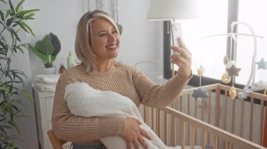 Sarışın genç kadın kundaklanmış bebeği ahşap karyolada tutuyor. Stüdyoda akıllı telefona gülümsüyor..