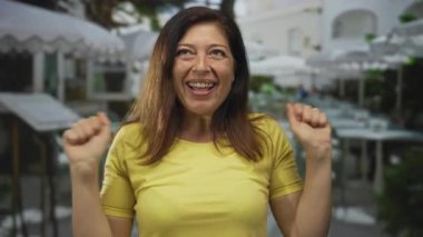 Orta yaşlı, sarı tişörtlü İspanyol kadın sokakta yumruklarını sıkar ve gülümser. Mutluluk..