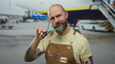 Havaalanı terminalinde makasıyla poz veren sakallı yakışıklı kel bir adam, açık havada stilist önlük giyerken açık uçuşu öneriyor..