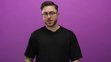 Stüdyo ortamında canlı mor bir arka planda, sakallı ve gözlüklü bir adam güvenle gülümsüyor, gençlik tavırları ve günlük siyah giysiler sergiliyor..