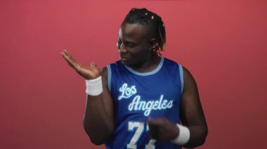 Adam, mavi basketbol forması ve beyaz bileklik giyerek pembe stüdyoda avucunun içini işaret ediyor..