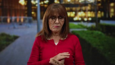 Gözlüklü son sınıf öğrencisi bir kadın şehir sokaklarında dikiliyor, kırmızı bir kazak giyiyor ve elleriyle işaret ediyor..