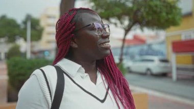 Açık havada örülmüş saçlarıyla gülümseyen kadın arka planda yeşillik ve modern mimari olan canlı şehir ortamını sergiliyor..