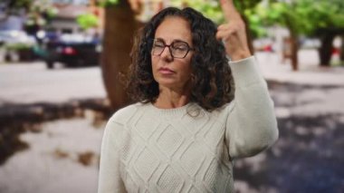 Kıvırcık saçlı, gözlüklü bir kadın şehir sokaklarında bir şeyin farkına varıyor. Düşünceli bir süveter giyiyor..