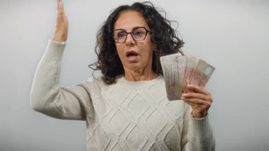 Orta yaşlı bir kadın, beyaz bir duvarın karşısında para sergiliyor. Şaşırdığını ve finansal açıdan başarılı olduğunu söylüyor..