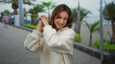 Güneşli bir sahil geçidinde neşeli bir şekilde gülümseyen, rahat bir kapüşonlu giyen, palmiye ağaçlarıyla çevrili, mutlu titreşimler gösteren ve rahatlamış açık havada keyif veren bir kadın..