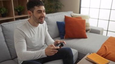Genç adam rahat bir oturma odasında rahat bir kanepede video oyunlarından hoşlanıyor. Çağdaş dekorları ve canlı yastıkları olan rahat bir ortam yaratıyor..