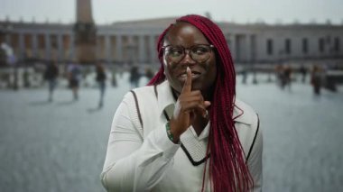 Gözlüklü ve kırmızı örgülü kadın, Vatikan 'ın Aziz Peter Meydanı' nda sessizlik için jestler yapıyor..