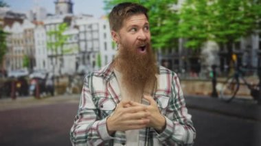 Sakallı ve küpeli bir adam şehir binalarında ağzı açık bir şekilde ellerini göğsüne yapıştırıyor..