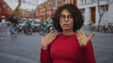 Kadın, park halindeki bisikletleri ve tuğlalı binaları olan, kırmızı süveter ve gözlük takan bir şehir caddesinde elleriyle hava üflüyor..