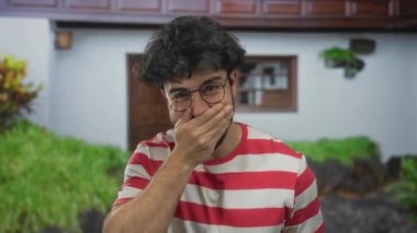 Güneşli bir günde açık havada gülümseyen sakallı ve gözlüklü genç bir adam yeşil çizgili bir gömlek ve arka planda bir ev giyiyor..