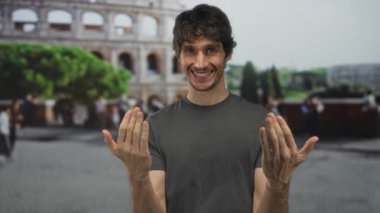Gülümseyen bir adam kolezyum binasının önünde elleri ile işaret eder; gezici keşif mirası mutluluğu.