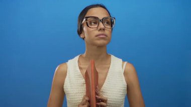 Ciddi bir ifadeyle izole edilmiş mavi arka planda kitap tutan, gözlüklü, beyaz bluzlu düşünceli ve çalışkan bir kadın.