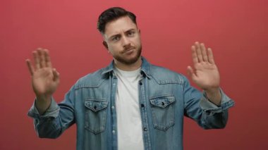 Kot gömlekli sakallı genç adam ellerini kırmızı arka plan duvarına dayayıp belirsizlik gösteriyor..