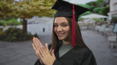 Kafkasyalı genç kız mezuniyet şapkası takıyor kırmızı püsküllü ve cüppeli gün ışığında el çırpıyor..