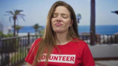 Kırmızı t-shirt giyen gönüllü kadın, deniz kenarındaki korkulukların yanında ellerini çeneye koyar; endişe yansıması.