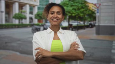 Binaların ve ağaçların yakınındaki bir caddede kollarını kavuşturmuş ve gülümseyen kadın, elleri kavuşturulmuş, güven güçlendirme pozitifliği.