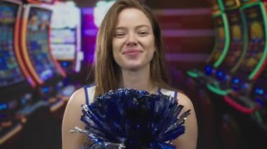 Genç bir kadın kumarhane binasında kumar makinesinin önünde tezahürat yaparken mavi ve gümüş ponponu sallıyor..