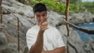 Deniz kenarında bulanık bir geçmişi olan genç bir adam güneşli bir günde etrafı ağaçlarla ve taşlarla çevrili beyaz bir gömlek giymiş düşünceli ve rahat görünüyor.