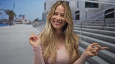 Pembe bluzlu sarışın kadın, beton basamaklı ve metal parmaklıklı deniz kenarında sağ ve sol parmaklarını işaret ediyor..