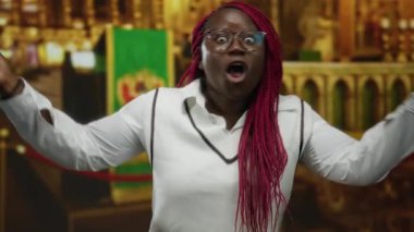 Gözlüklü ve kırmızı örgülü bir kadın kapalı bir kilise ortamında anlamlı bir şekilde konuşur, duygularını aktarır ya da şaşırtır..