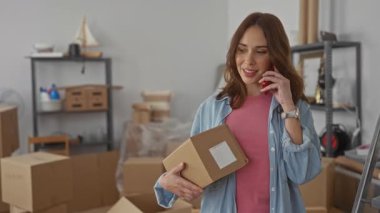 Yeni evdeki telefon kulübesinde konuşan bir kadın taşınma gününün rahat atmosferini gösteren bir apartmanın oturma odasında kutular ve raflarla çevrili..