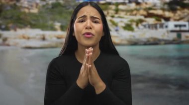 Kadın ellerini tutup plajda yalvarmaya başlıyor. Deniz kenarında yardım dilenen Latin kökenli bir genç figürle..