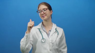 Beyaz önlüklü, steteskop ve gözlüklü genç sarışın kadın doktor mavi bir stüdyoda parmağını omzunun üstünden işaret ediyor..