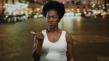 Beyaz atletli bir kadın gecenin bir yarısı hareketli bir caddede duruyor, sokak lambalarının altında düşünceli bir ifadeyle kenarı işaret ediyor..