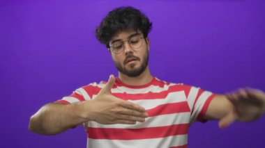 Sakallı genç adam mor arka plana karşı çerçeve hareketi yapıyor. Yaratıcılık taslıyor ve çizgili bir gömlek giyiyor. Hareketli bir hava yaratıyor..