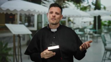 Rahip terasta duruyor, papaz cübbesi ve haç giyiyor, elinde bir İncil tutuyor, açık hava restoranının arka planına sahip bir dinginlik yayıyor, ruhani tefekkür sunuyor..