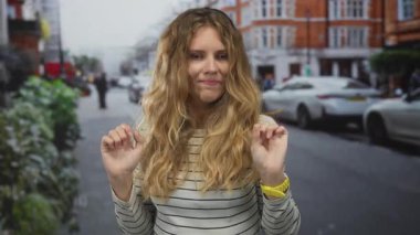 Meşgul bir caddede, hareketsiz bir şekilde el kaldıran kadın, sarışın endişeli bakışları, açık hava fotoğrafındaki dalgalı saçlarla çerçevelenmiş..
