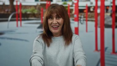 Kıdemli İspanyol kadın gülümsüyor ve elleri açık bir spor salonuna gelmek için işaret ediyor. Etrafı parlak kırmızı egzersiz ekipmanlarıyla çevrili, sıcaklık ve enerji taşıyor..