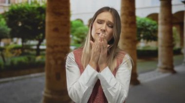 Şehir sokaklarında üzgün görünen bir kadın pembe süveter giyiyor. Arka planda sütunlar ve yeşillikler var. Şehir ortamında duygusal bir sahne olduğunu gösteriyor..