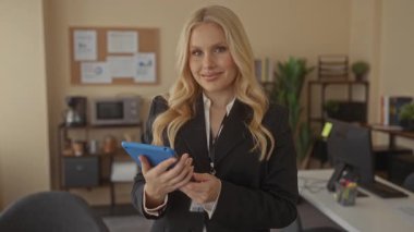 Ofiste elinde tablet tutan, gülümseyen sarı saçlı profesyonel bir kadın geniş bir çalışma alanına benziyor..