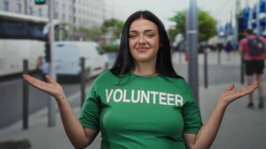 Şehir caddesinde gülümseyen, yeşil tişörtlü, otobüslerle çevrili ve açık hava ortamında yaşayan genç ve iri yarı bir kadın gönüllü..