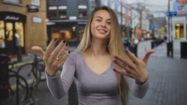 Arka planda canlı binalar olan bir şehir caddesinde davetkar bir şekilde el kol hareketi yapan bir kadın, kentsel bir ortamda dostane ve misafirperver bir atmosfer taşıyor..