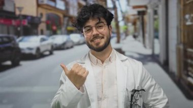 Young bearded hispanic man wearing glasses and a white coat smiles confidently outdoors by the street, giving a thumbs-up gesture, conveying positivity and assurance.