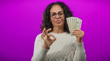 Gözlüklü kadın, canlı mor bir arka plana karşı iyi bir jest yaparak bize dolar veriyor, kendine güven ve başarı gösteriyor..