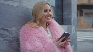 Açık havada konuşan, pembe ceketli, sarı saçlı şehir duvarına yaslanmış bir kadın..